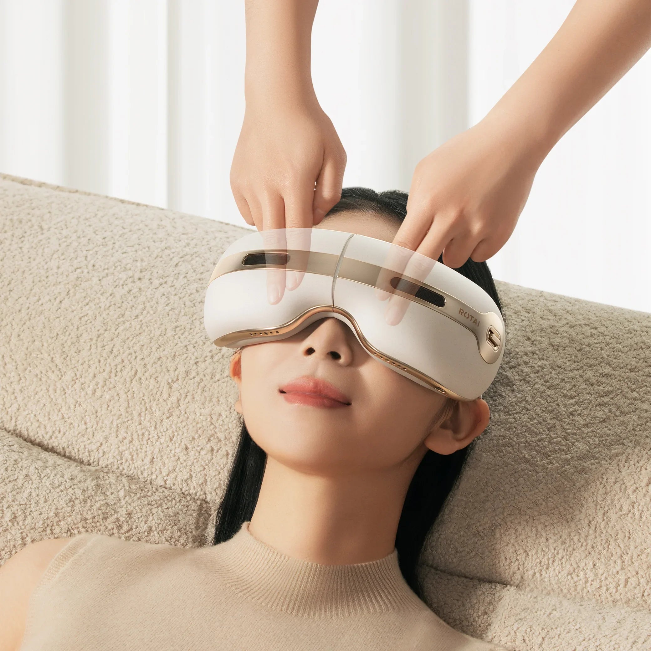 Woman relaxing with white and gold eye massager device indoors on beige couch