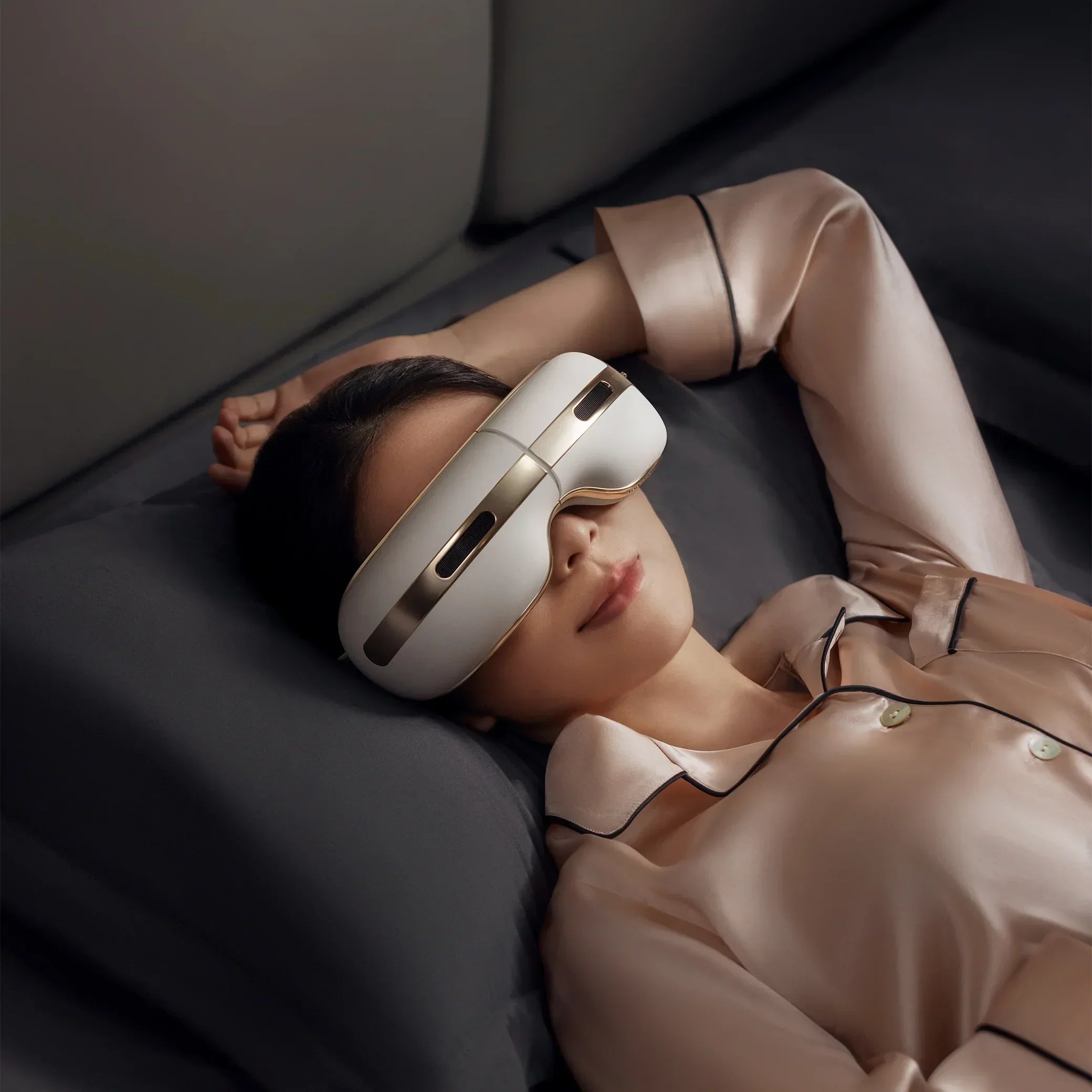 Woman lying on bed wearing beige silk pajamas using a white and gold eye massager device
