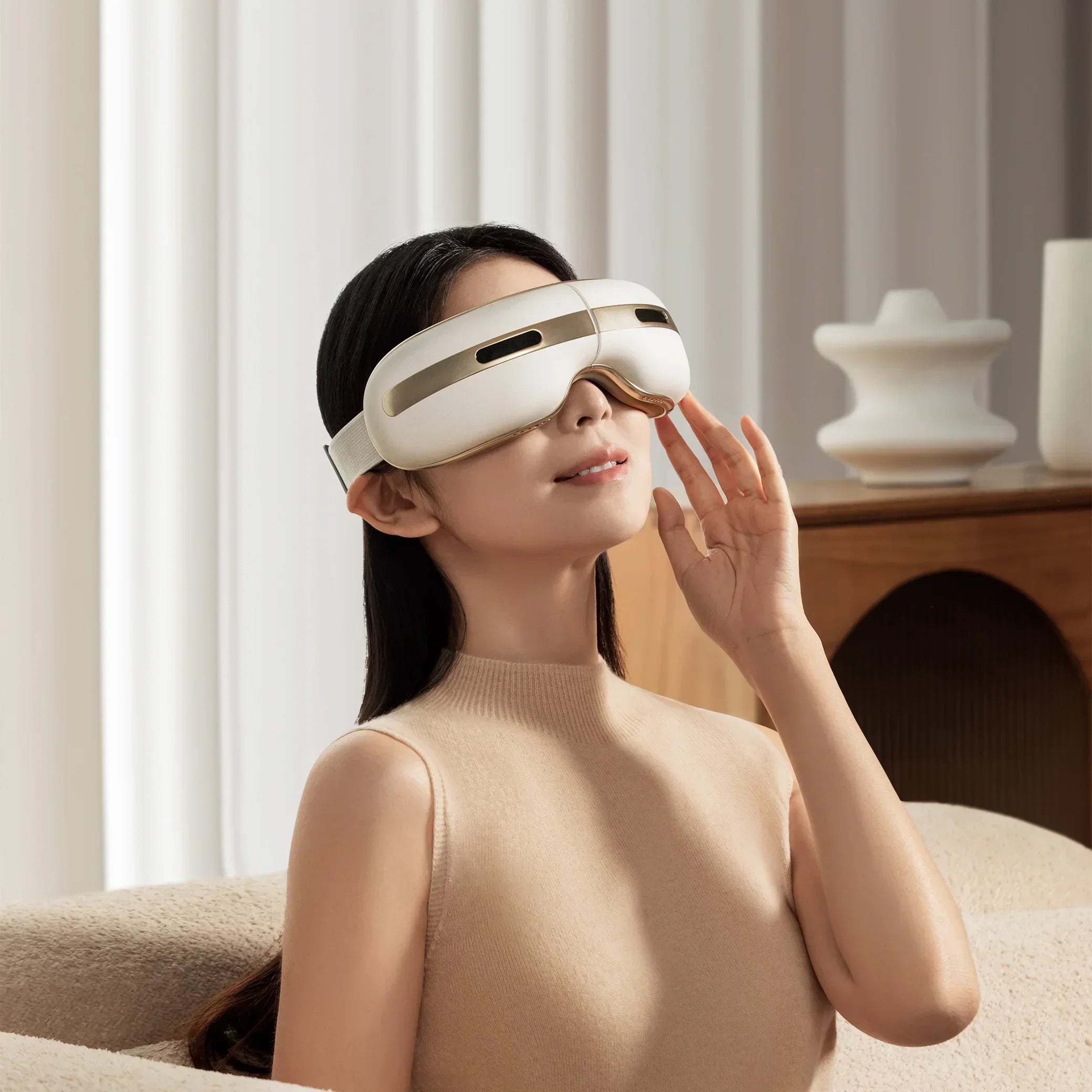 Woman wearing white and gold eye massager sitting in modern beige living room