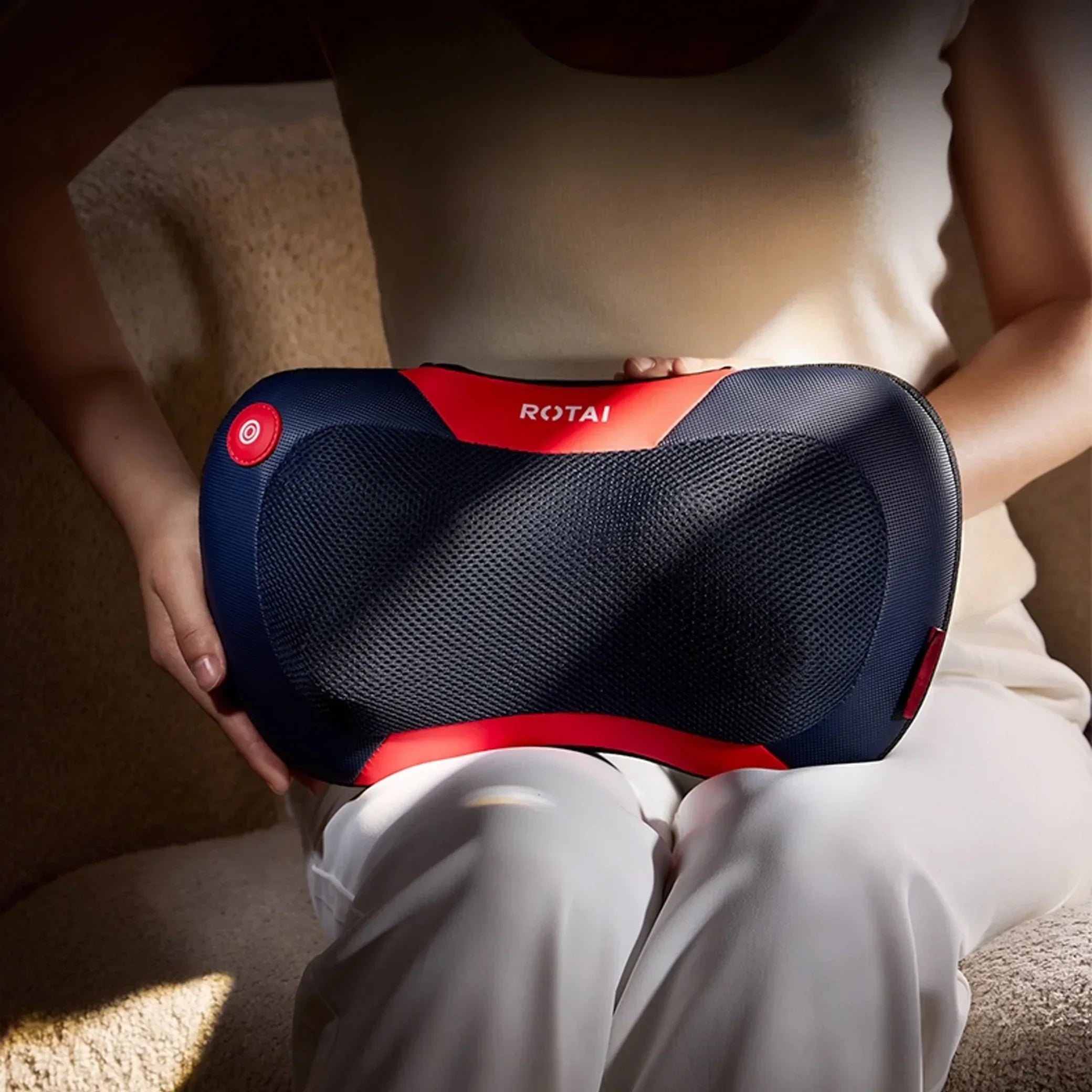 Person holding ROTAI K30 massager pillow in lap on beige couch, black mesh and red trim