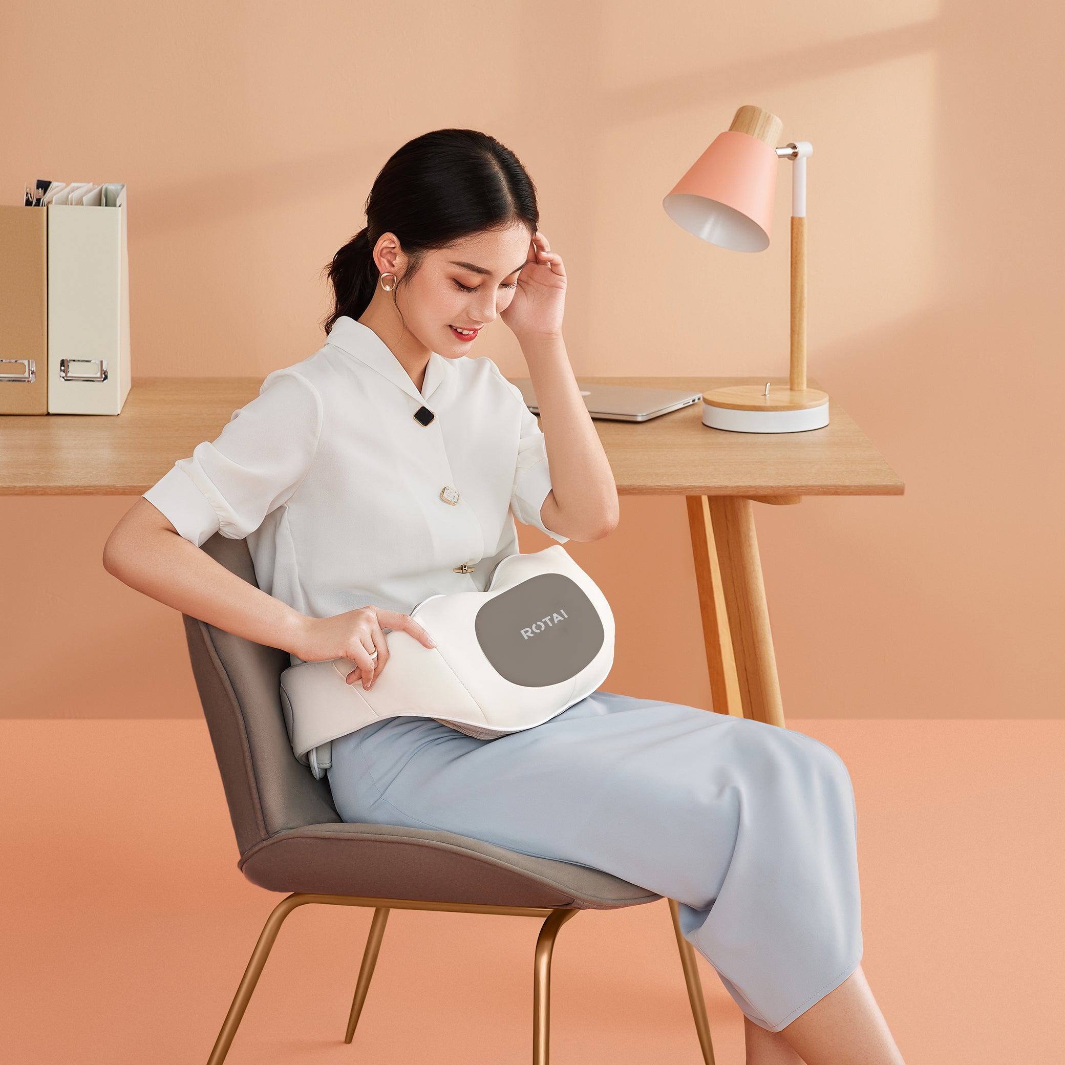 Woman sitting on chair using ROTAI white massage belt in modern peach-colored workspace