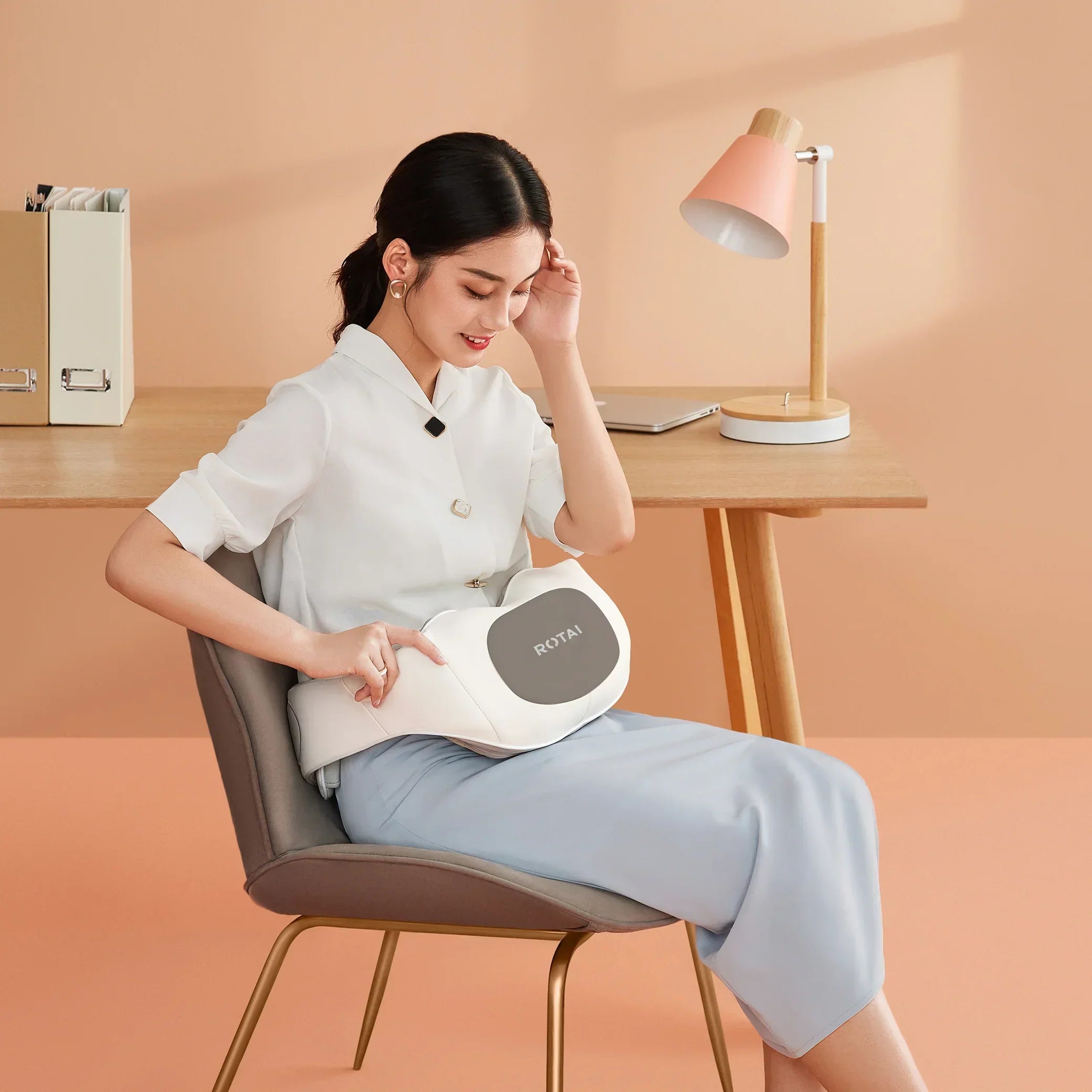Woman sitting on chair using ROTAI white massage belt in modern peach-colored workspace