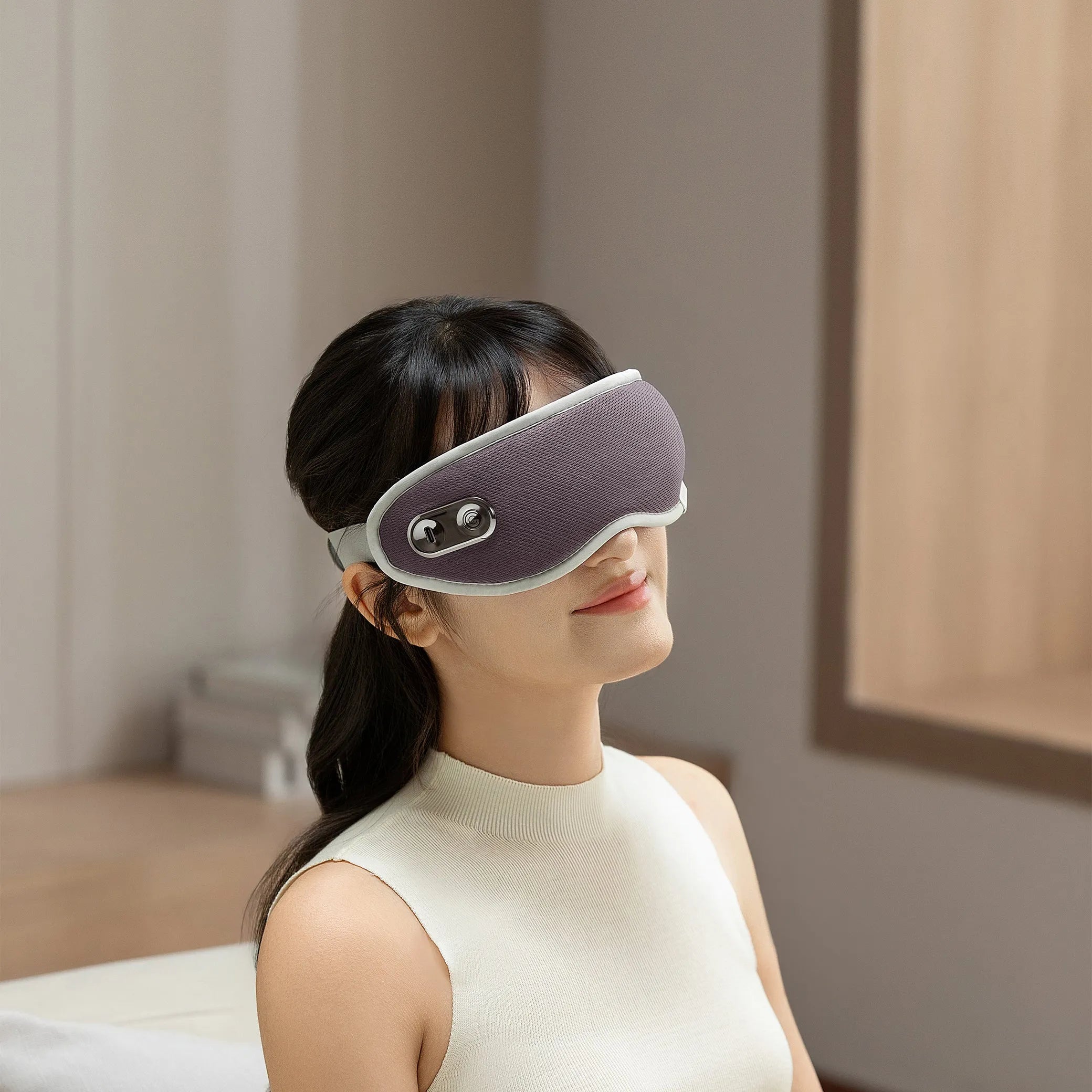 Woman wearing a lavender eye massager mask in a softly lit modern room