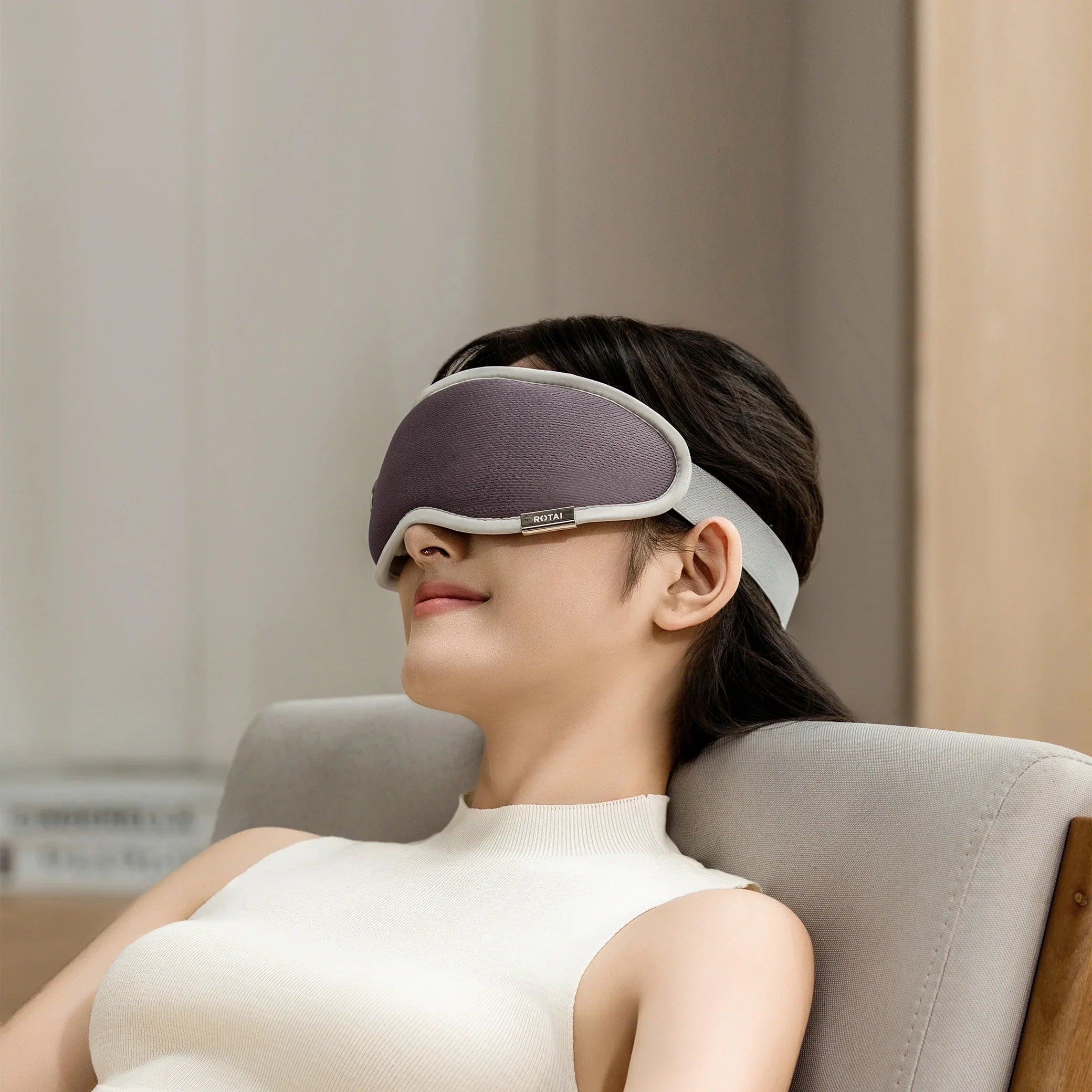 Woman relaxing on a chair wearing a gray eye mask with white trim in a cozy indoor setting