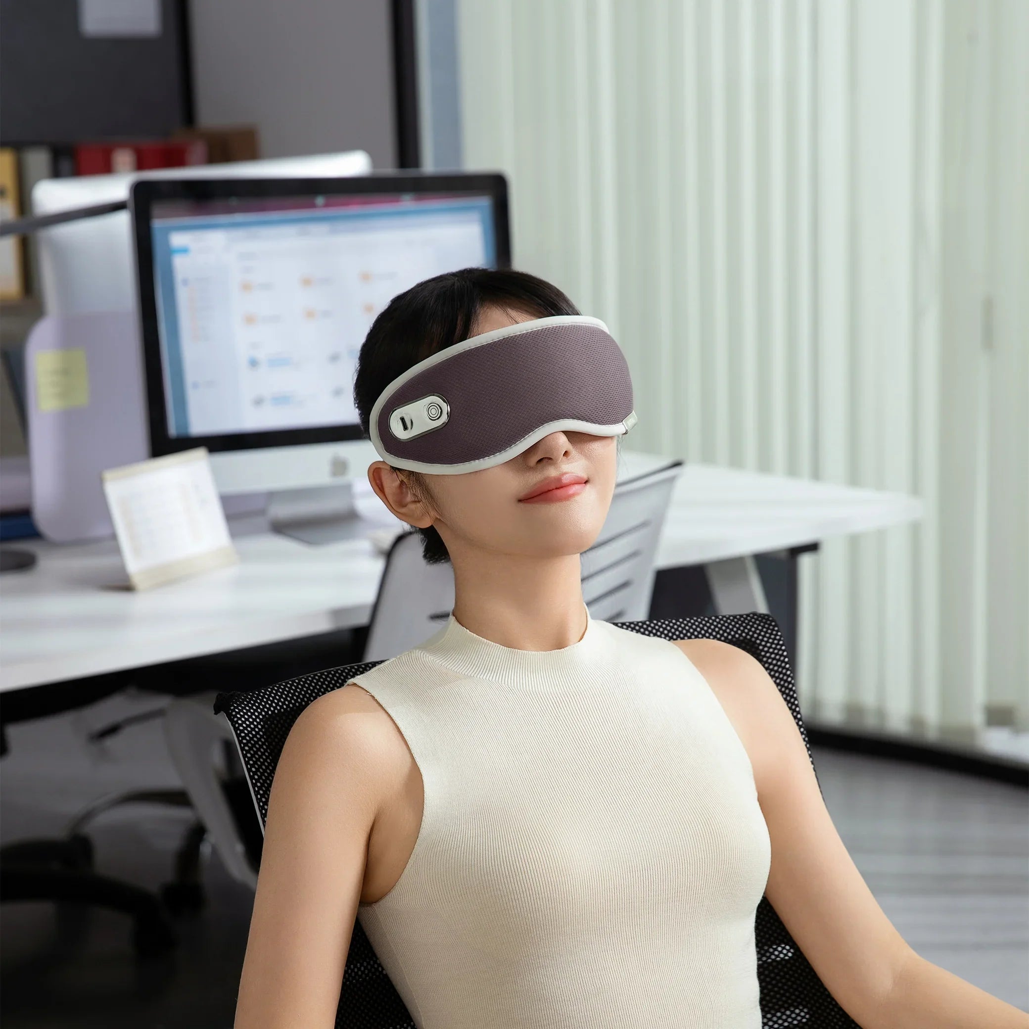 Woman wearing a gray eye massager relaxing in modern office chair at desk with computer