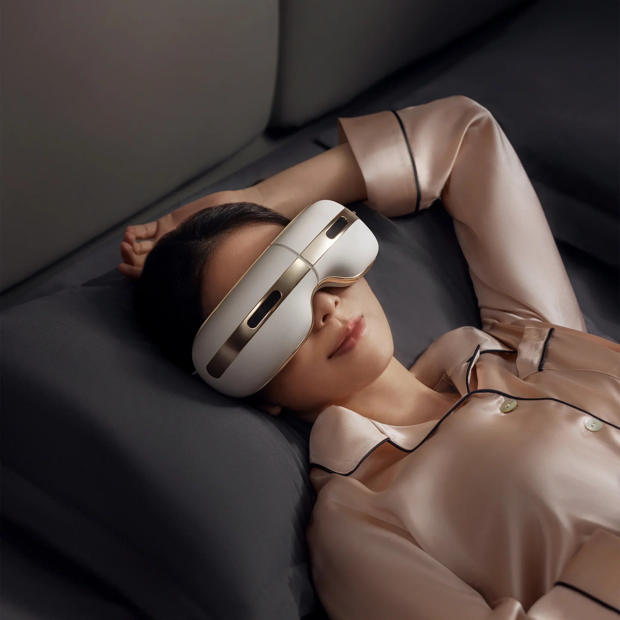 Woman lying on bed wearing beige silk pajamas using a white and gold eye massager device