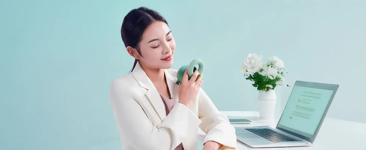 Woman using a green scalp massager on shoulder at desk with laptop and flowers